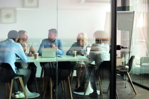 Image includes a view of five people gathered around a table in an office, with coffees and a laptop having a conversation