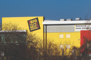 A bright yellow building with a sign reading "The Big Yellow Self Storage Company" on the side, partially obscured by leafless trees in the foreground. Another sign on the right side of the building reads "Business & home storage" against a clear blue sky.